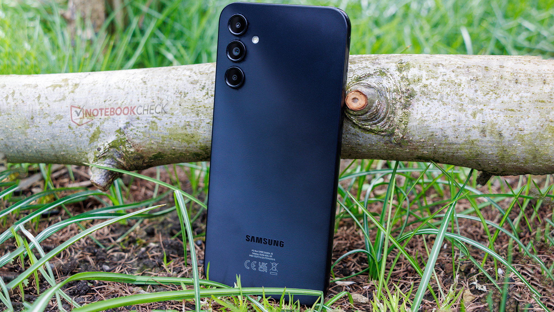  The image shows the back of a black Samsung Galaxy A14 5G smartphone with a 50 MP camera, lying on a wooden log outdoors.