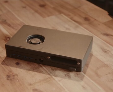 Una consola de videojuegos negra y compacta con una unidad de disco integrada, puertos USB y un ventilador de refrigeración situado en la parte superior, apoyada sobre una mesa de madera.