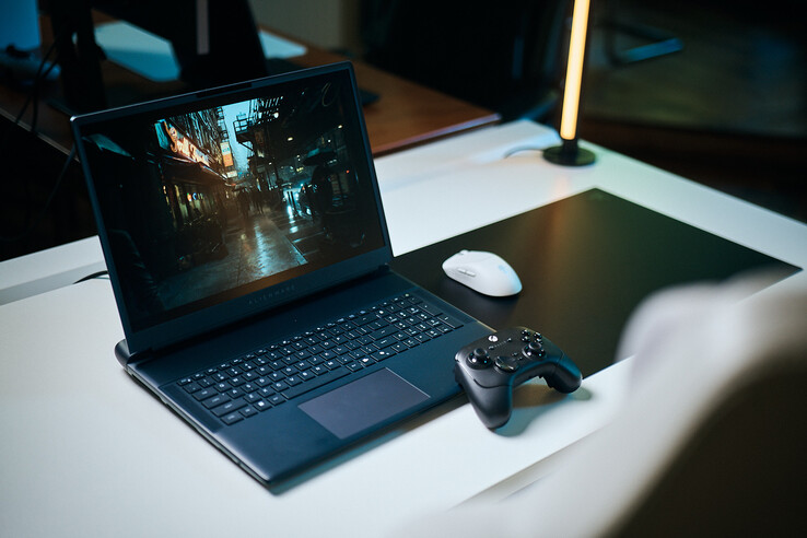 Un ordenador portátil para videojuegos Alienware abierto, colocado sobre un escritorio blanco de estudio, en el que se muestra una escena urbana oscura y reflectante con un nítido nivel de detalle obtenido mediante el método de trazado de trayectorias, acompañado de un mando negro y un ratón inalámbrico blanco.