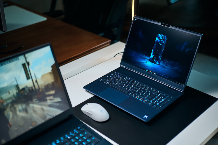 Un portátil para juegos de gama media de color azul oscuro (Alienware Aurora 16X) colocado en diagonal sobre una alfombrilla de escritorio con un fondo de pantalla de ciencia ficción azul brillante, situado junto a un ratón inalámbrico blanco, con otro portátil difuminado en primer plano.