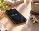 Un smartphone que muestra una forma de onda de audio junto a unos auriculares inalámbricos y una taza de café sobre un escritorio de madera
