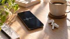 Un smartphone que muestra una forma de onda de audio junto a unos auriculares inalámbricos y una taza de café sobre un escritorio de madera