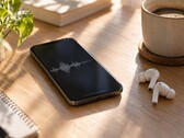 Un smartphone que muestra una forma de onda de audio junto a unos auriculares inalámbricos y una taza de café sobre un escritorio de madera