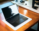 Un portátil para juegos Razer Blade 16 de color negro mate, con la tapa abierta, colocado sobre una alfombrilla de escritorio blanca que recubre una mesa de madera de tonos cálidos, en el que se muestra un fondo de pantalla en blanco y negro de gran intensidad.