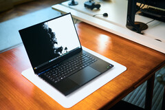Un portátil para juegos Razer Blade 16 de color negro mate, con la tapa abierta, colocado sobre una alfombrilla de escritorio blanca que recubre una mesa de madera de tonos cálidos, en el que se muestra un fondo de pantalla en blanco y negro de gran intensidad.
