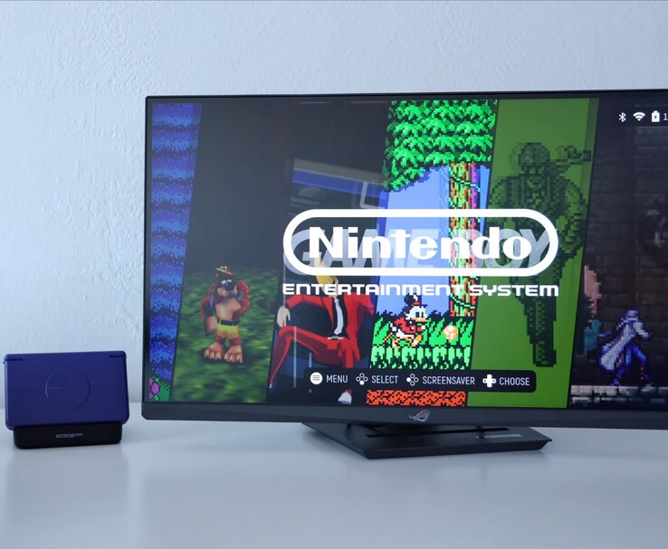 Retroid Pocket Flip 2 en color púrpura colocado en un dock junto a un monitor, con la pantalla del televisor mostrando una interfaz de emulador retro de NES. (Fuente de la imagen: Retro Game Corps en YouTube)