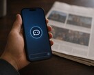Una mano sostiene un smartphone en el que se ve el icono de un chatbot; junto a él hay un periódico impreso.