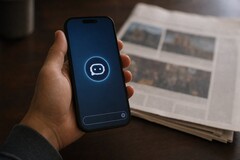 Una mano sostiene un smartphone en el que se ve el icono de un chatbot; junto a él hay un periódico impreso.