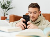Un adolescente con sudadera con capucha escribiendo en un smartphone con los libros de clase abiertos delante de él