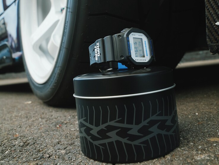 Reloj Casio G-Shock DW5600TT25-1 x Toyo Tires expuesto en una caja metálica con motivos de neumáticos, junto a una rueda de coche.
