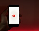 Una mano que sostiene un smartphone con el logotipo de YouTube iluminado en la pantalla sobre un fondo rojo