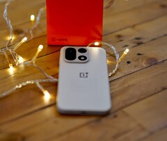 Un OnePlus 15 descansa sobre una mesa de madera rodeado de cálidas guirnaldas de luces, con una caja de venta al público de OnePlus de color rojo brillante situada al fondo.