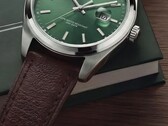 Primer plano del reloj Timex Legacy Day and Date, que muestra su esfera verde con acabado de rayos de sol, bisel de tono plateado y asas cepilladas, corona estriada y correa de cuero marrón texturizado sobre una caja verde oscura y una superficie de madera.