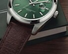 Primer plano del reloj Timex Legacy Day and Date, que muestra su esfera verde con acabado de rayos de sol, bisel de tono plateado y asas cepilladas, corona estriada y correa de cuero marrón texturizado sobre una caja verde oscura y una superficie de madera.