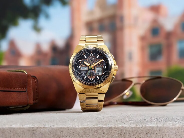 Reloj de piloto Citizen Promaster Skyhawk A-T JY8144-50E en tono dorado, expuesto en posición vertical sobre una repisa junto a un bolso de piel marrón y unas gafas de sol, con un paisaje urbano difuminado al fondo.
