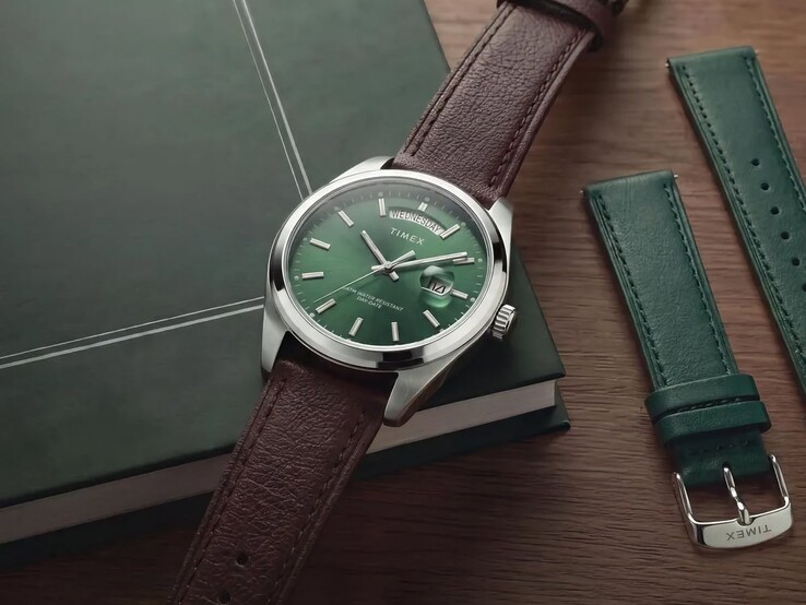 El Timex Legacy Day and Date con su esfera verde y correa de cuero marrón, colocado sobre un libro verde oscuro junto a la correa intercambiable de cuero verde.