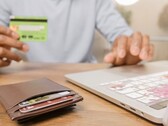 Persona sosteniendo una tarjeta bancaria frente a un portátil, con una cartera sobre el escritorio