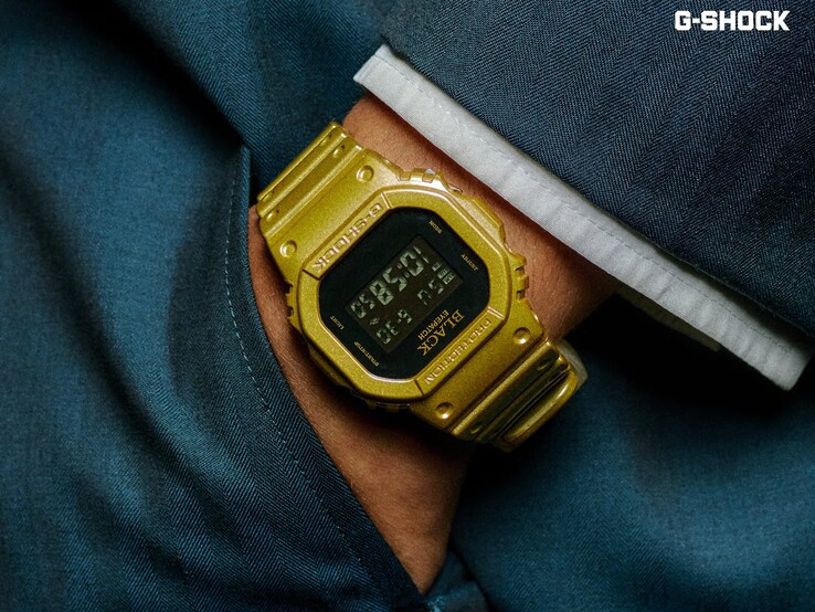 Primer plano de un reloj Casio G-Shock DW-5600BEP-9 dorado que se lleva en la muñeca sobre un tejido azul oscuro. El reloj cuenta con una pantalla digital negra, caja y correa doradas, y el logotipo de BlackEyePatch en el bisel y la esfera.