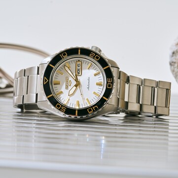 Reloj Seiko 5 Sports SKX HDB004 con esfera blanca plateada, bisel giratorio negro con marcas de color dorado, agujas e índices dorados y brazalete de acero inoxidable, fotografiado en ángulo.