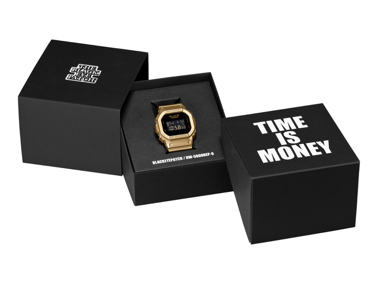 Un reloj Casio G-Shock DW-5600BEP-9 dorado con pantalla digital negra se encuentra entre dos estuches de presentación negros. El reloj presenta la marca BlackEyePatch en la esfera, mientras que en el estuche del primer plano se lee «TIME IS MONEY».