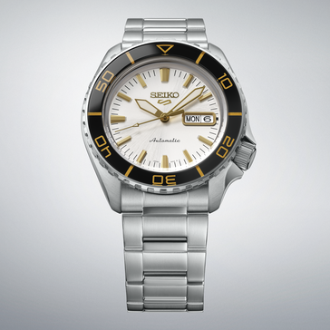 Seiko 5 Sports SKX HDB004 con esfera blanca plateada, bisel negro con marcas de color dorado, agujas e índices en tono dorado y brazalete de acero inoxidable, mostrado sobre un fondo gris claro.